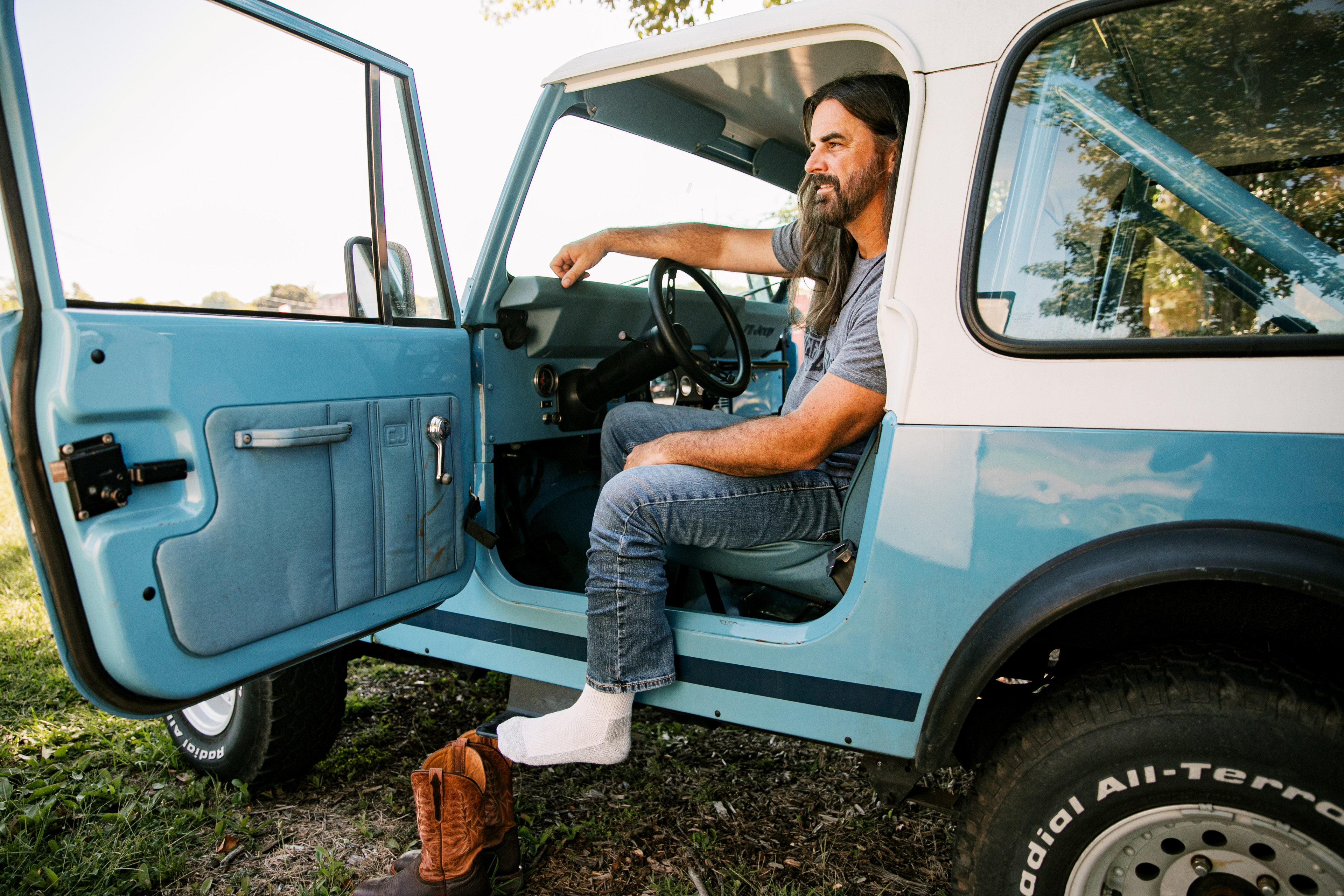 Man sitting inside a vintage blue and white van with open door, surrounded by greenery.
