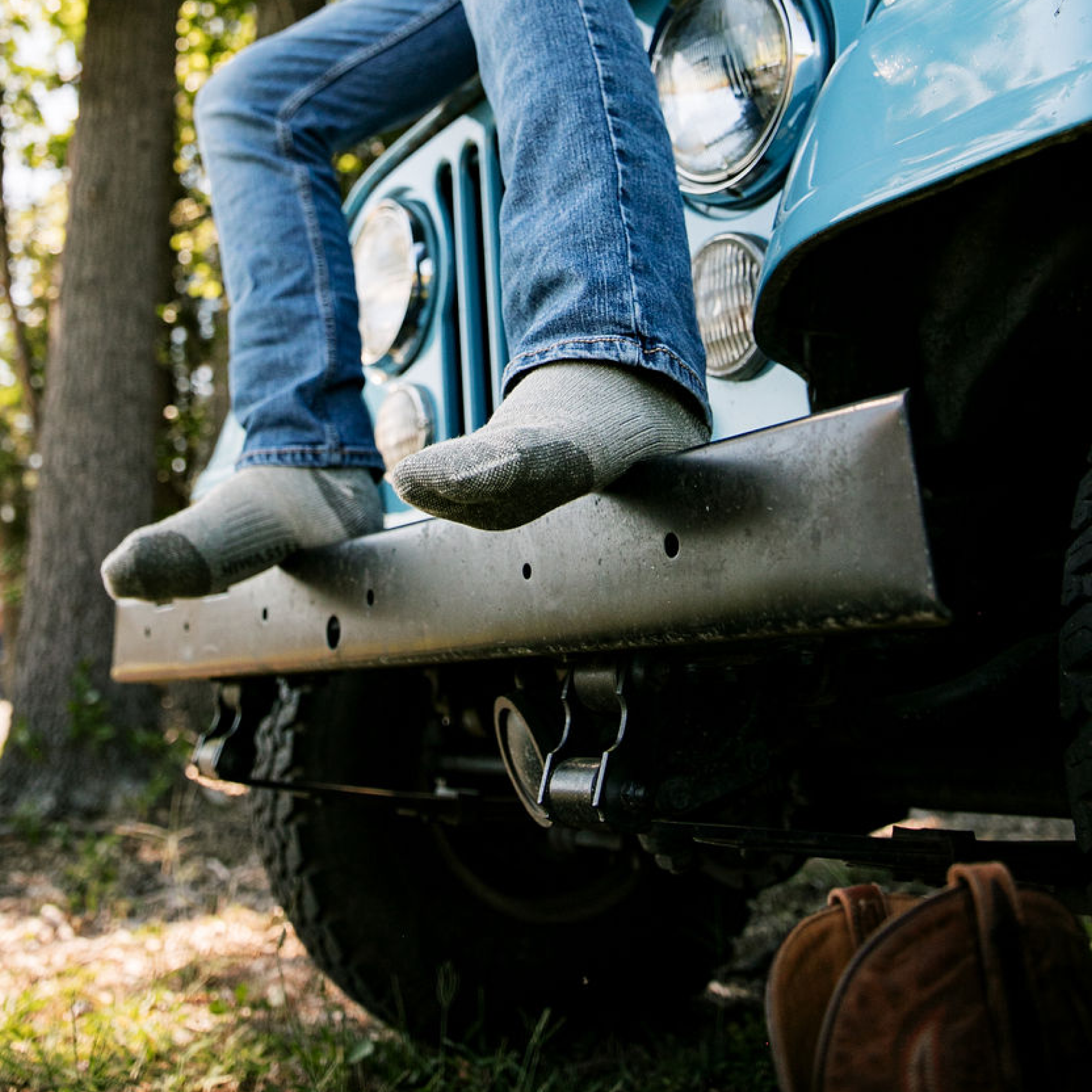 A person wearing blue jeans and gray socks sits on the front bumper of a vintage blue jeep outdoors in a wooded setting.